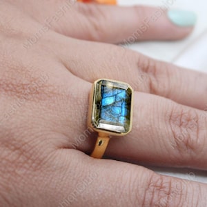 May include: A gold ring featuring a large, rectangular, iridescent blue labradorite gemstone. The ring is set in a plain, gold band.