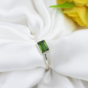 May include: A silver ring with a rectangular, emerald-green gemstone. The ring is set against white fabric, with yellow flowers in the background. The gemstone is approximately 0.8 cm in length.