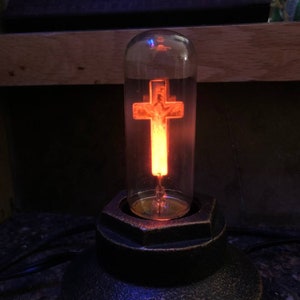 May include: A clear glass vacuum tube light bulb containing a glowing orange cross filament. The bulb is mounted in a dark, textured, hexagonal metal base. The orange light illuminates the interior of the glass cylinder, creating a warm glow.