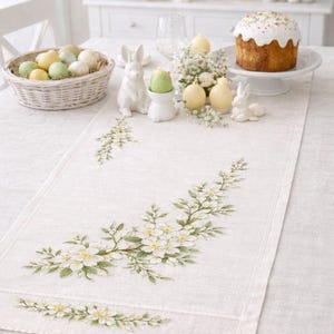 Può includere: Runner da tavolo in lino bianco con ricamo floreale. Il runner presenta un delicato motivo di fiori bianchi e foglie verdi. La tavola è apparecchiata per Pasqua con uova decorative, coniglietti e una torta.