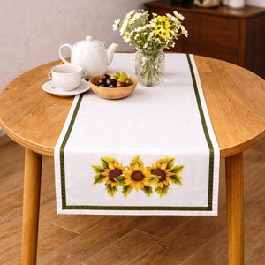 Könnte beinhalten: Weißer Tischläufer mit grünem Rand und gestickten Sonnenblumen. Der Läufer liegt auf einem runden Holztisch, mit einer Teekanne, einer Tasse, einer Schale mit Trauben und einer Vase mit Blumen.