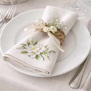Puede incluir: Una servilleta de lino blanco doblada con un diseño floral bordado, acentuada con hojas verdes y centros amarillos, descansa sobre un plato blanco. Un servilletero tejido con pequeñas flores blancas y una cinta crema adorna la servilleta. Cubiertos y una cesta de huevos en el fondo.