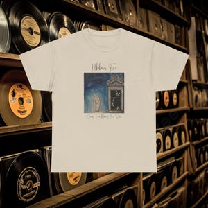 May include: A beige t-shirt with a graphic print of a painting. The painting depicts two figures standing in front of a building with a clock. The text "Ribbon Fix" and "Camp Fire Does The Sun" are printed on the shirt.