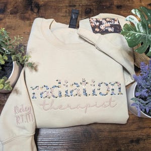 May include: A beige sweatshirt with floral embroidered text that reads "radiation therapist." The sleeve has the embroidered text "Rickey R.T.(T)." The sweatshirt is folded on a wooden surface, with plants in the background.