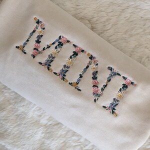 K&ouml;nnte beinhalten: Cremefarbenes Kissen mit dem gestickten Wort "NANA" in floralen Buchstaben. Die Stickerei zeigt rosa, blaue, gelbe und wei&szlig;e Blumen mit gr&uuml;nen Bl&auml;ttern. Das Kissen liegt auf einer wei&szlig;en, flauschigen Oberfl&auml;che.