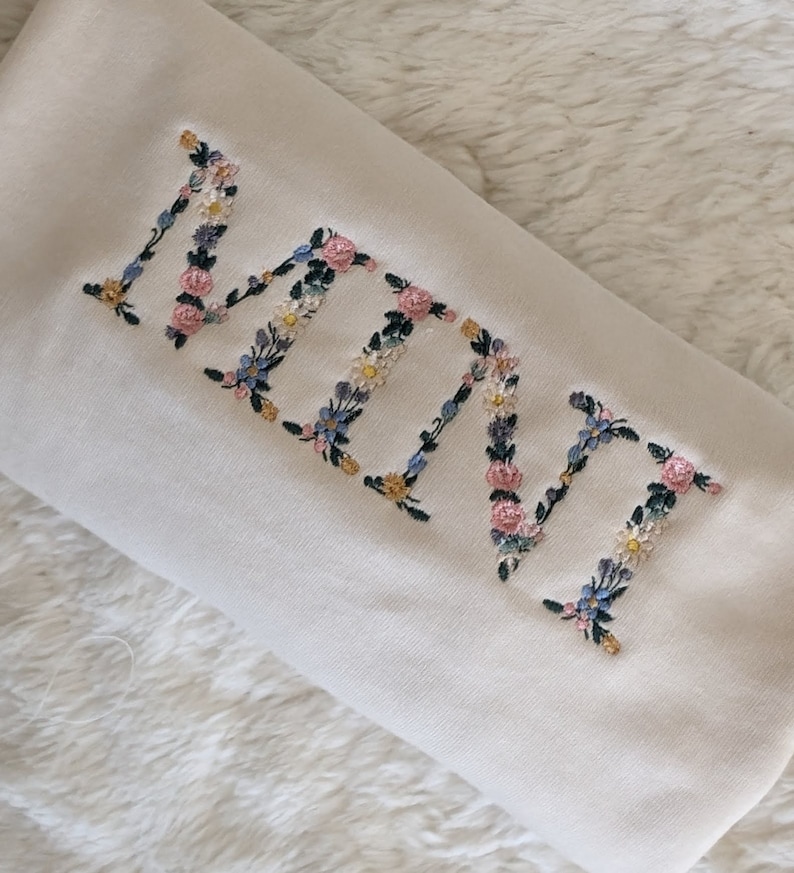 K&ouml;nnte beinhalten: Cremefarbenes Kissen mit dem gestickten Wort "NANA" in floralen Buchstaben. Die Stickerei zeigt rosa, blaue, gelbe und wei&szlig;e Blumen mit gr&uuml;nen Bl&auml;ttern. Das Kissen liegt auf einer wei&szlig;en, flauschigen Oberfl&auml;che.