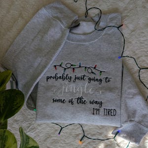 Pu&ograve; includere: Felpa grigio chiaro con la scritta ricamata "probably just going to jingle some of the way I'M TIRED". Il design include una stringa di luci natalizie colorate sopra il testo.