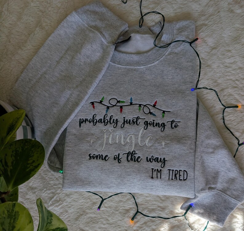Pu&ograve; includere: Felpa grigio chiaro con la scritta ricamata "probably just going to jingle some of the way I'M TIRED". Il design include una stringa di luci natalizie colorate sopra il testo.