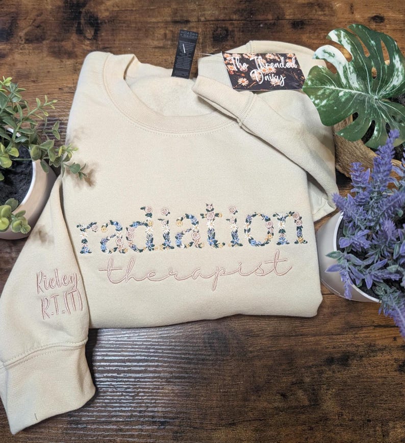 May include: A beige sweatshirt with floral embroidered text that reads "radiation therapist." The sleeve has the embroidered text "Rickey R.T.(T)." The sweatshirt is folded on a wooden surface, with plants in the background.