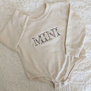 K&ouml;nnte beinhalten: Cremefarbener Baby-Body mit langen &Auml;rmeln. Das Wort "MINI" ist mit einem Blumenmuster mit rosa, blauen und gelben Blumen auf die Brust gestickt. Der Body hat Druckkn&ouml;pfe am unteren Ende.