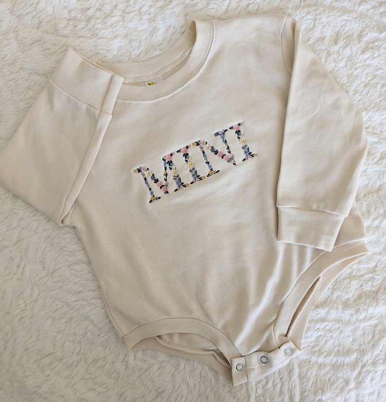 K&ouml;nnte beinhalten: Cremefarbener Baby-Body mit langen &Auml;rmeln. Das Wort "MINI" ist mit einem Blumenmuster mit rosa, blauen und gelben Blumen auf die Brust gestickt. Der Body hat Druckkn&ouml;pfe am unteren Ende.