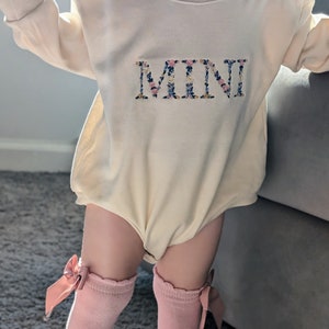 K&ouml;nnte beinhalten: Ein cremefarbener Baby-Strampler mit langen &Auml;rmeln und dem Wort "MINI" in einem Blumenmuster gestickt. Das Baby tr&auml;gt rosa kniehohe Socken mit Schleifen.