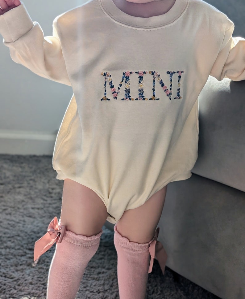 K&ouml;nnte beinhalten: Ein cremefarbener Baby-Strampler mit langen &Auml;rmeln und dem Wort "MINI" in einem Blumenmuster gestickt. Das Baby tr&auml;gt rosa kniehohe Socken mit Schleifen.