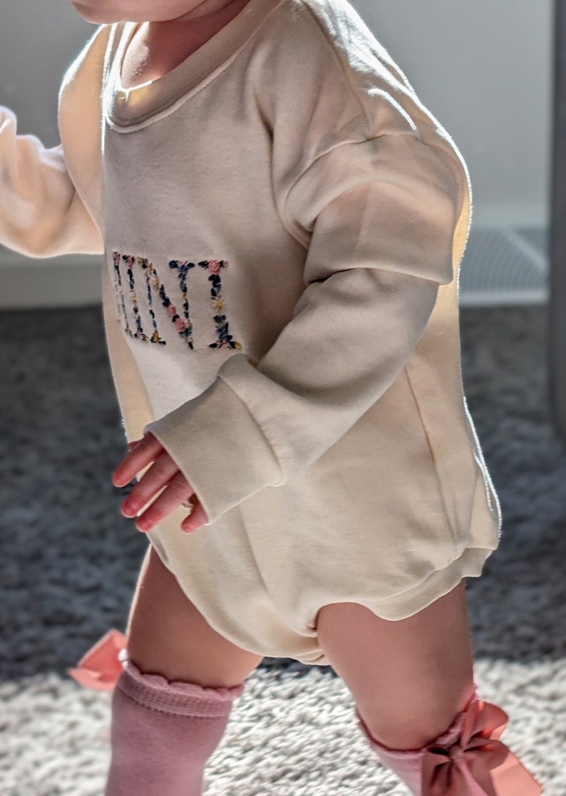 K&ouml;nnte beinhalten: Cremefarbener Baby-Strampler mit langen &Auml;rmeln. Das Wort "MINI" ist vorne in einem Blumenmuster gestickt. Das Baby tr&auml;gt au&szlig;erdem rosa Kniestr&uuml;mpfe mit Schleifen.