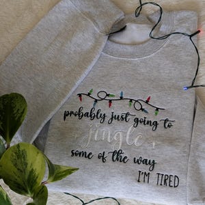 Pu&ograve; includere: Felpa grigio chiaro con la scritta ricamata "probably just going to jingle some of the way I'M TIRED". Il design include una fila di luci natalizie colorate. La felpa &egrave; piegata.