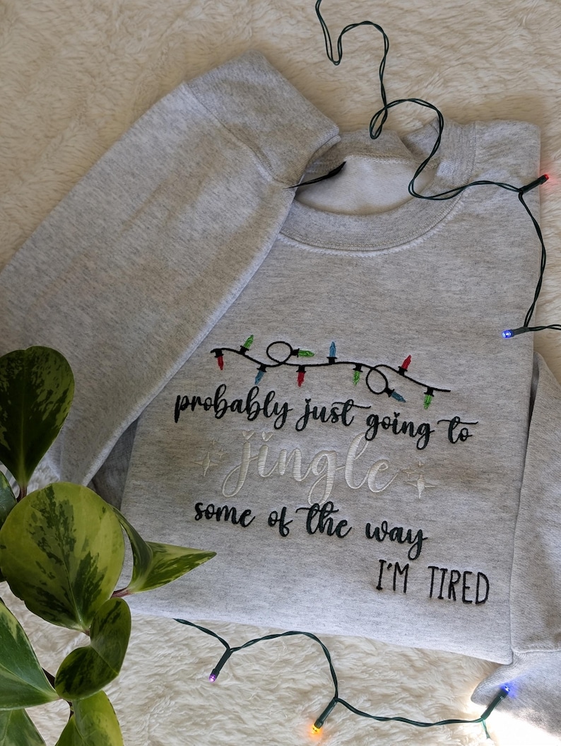 Pu&ograve; includere: Felpa grigio chiaro con la scritta ricamata "probably just going to jingle some of the way I'M TIRED". Il design include una fila di luci natalizie colorate. La felpa &egrave; piegata.
