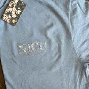 K&ouml;nnte beinhalten: Ein hellblaues T-Shirt mit den Worten "NICU nurse", die in Wei&szlig; mit kleinen Blumen um die Buchstaben gestickt sind.