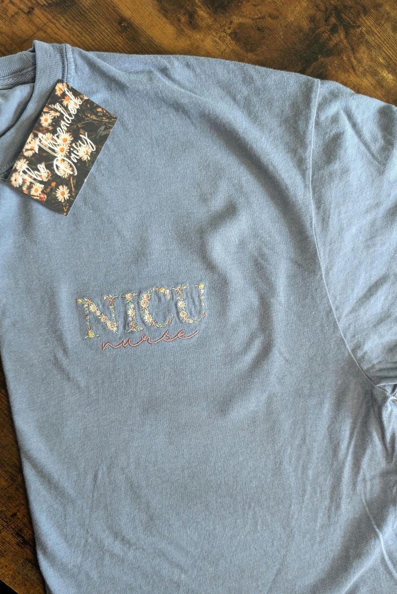 K&ouml;nnte beinhalten: Ein hellblaues T-Shirt mit den Worten "NICU nurse", die in Wei&szlig; mit kleinen Blumen um die Buchstaben gestickt sind.