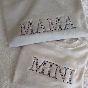 K&ouml;nnte beinhalten: Zwei Sweatshirts, eines hellbeige und eines cremefarben, mit floralen Buchstaben bestickt. Auf dem oberen Sweatshirt steht "MAMA" und auf dem unteren "MINI". Die Blumenstickerei umfasst rosa, blaue und gelbe Blumen.