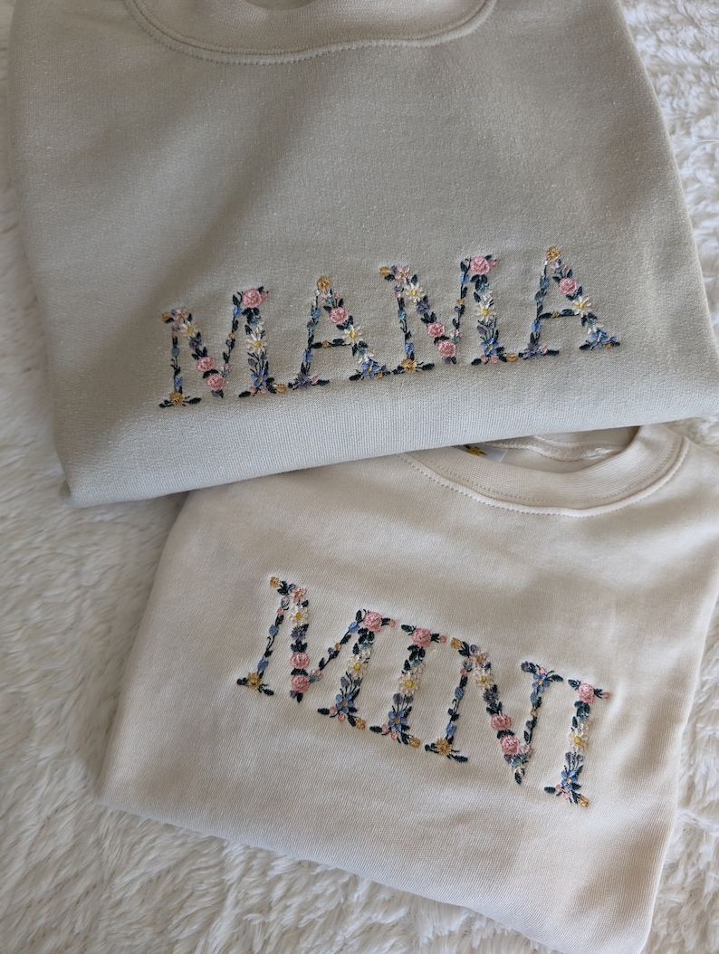 K&ouml;nnte beinhalten: Zwei Sweatshirts, eines hellbeige und eines cremefarben, mit floralen Buchstaben bestickt. Auf dem oberen Sweatshirt steht "MAMA" und auf dem unteren "MINI". Die Blumenstickerei umfasst rosa, blaue und gelbe Blumen.