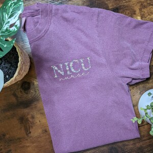 K&ouml;nnte beinhalten: Ein lila T-Shirt mit dem Text "NICU nurse" in Wei&szlig; mit floralen Akzenten.