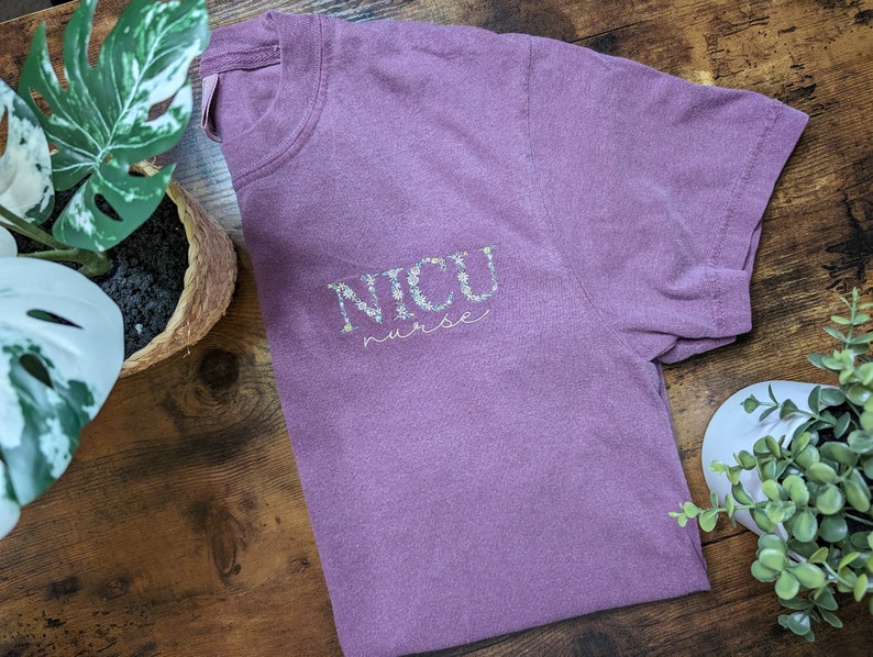 K&ouml;nnte beinhalten: Ein lila T-Shirt mit dem Text "NICU nurse" in Wei&szlig; mit floralen Akzenten.