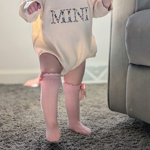 K&ouml;nnte beinhalten: Ein cremefarbener Baby-Strampler mit dem Wort "MINI" in floralen Buchstaben gestickt. Das Baby tr&auml;gt rosa kniehohe Socken mit Schleifen. Das Outfit ist f&uuml;r ein Baby konzipiert.