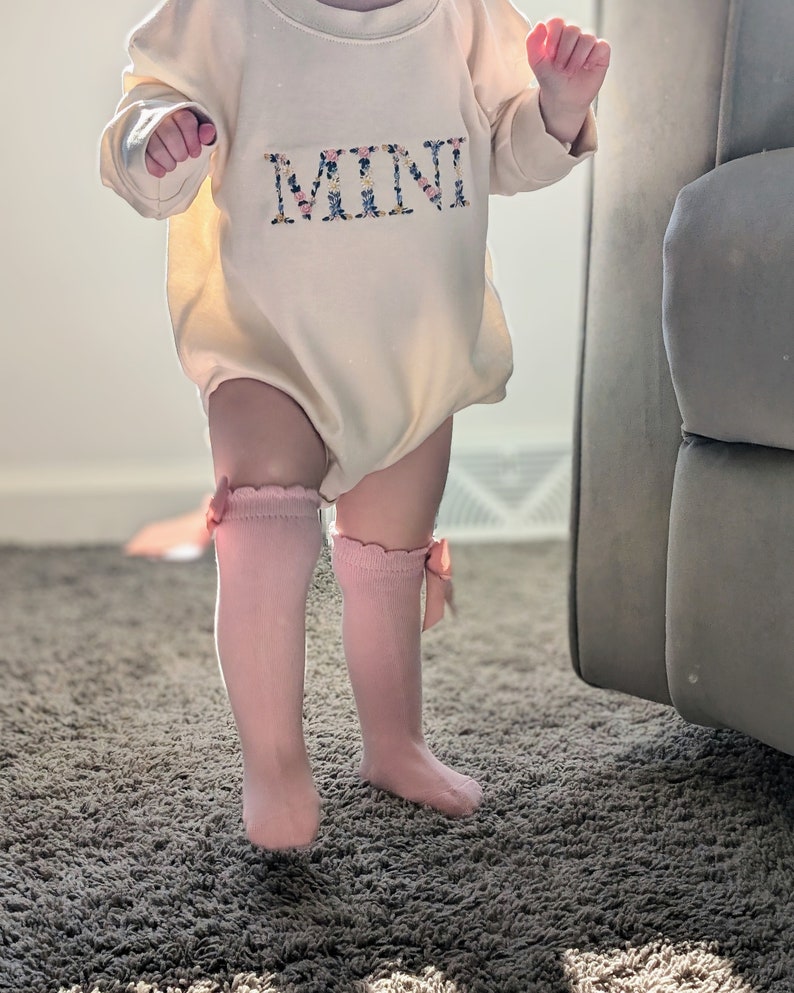 K&ouml;nnte beinhalten: Ein cremefarbener Baby-Strampler mit dem Wort "MINI" in floralen Buchstaben gestickt. Das Baby tr&auml;gt rosa kniehohe Socken mit Schleifen. Das Outfit ist f&uuml;r ein Baby konzipiert.
