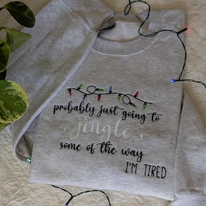 Pu&ograve; includere: Felpa grigio chiaro con la scritta ricamata "probably just going to jingle some of the way I'M TIRED". Il design include una stringa di luci natalizie colorate.