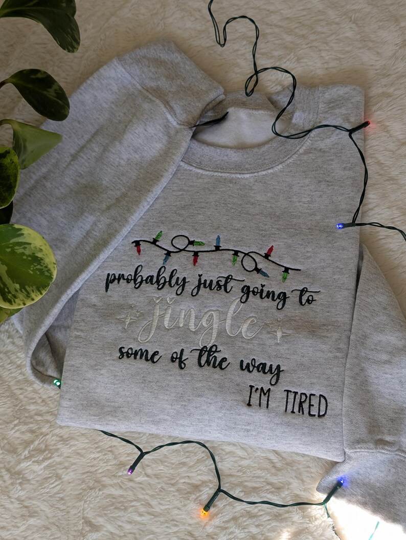 Pu&ograve; includere: Felpa grigio chiaro con la scritta ricamata "probably just going to jingle some of the way I'M TIRED". Il design include una stringa di luci natalizie colorate.