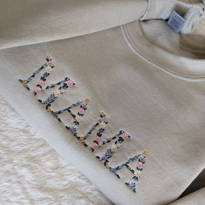 K&ouml;nnte beinhalten: Hellgr&uuml;nes Sweatshirt mit dem gestickten Wort "MAMA" in floralen Buchstaben. Die Buchstaben sind mit rosa Rosen, blauen und gelben Wildblumen und gr&uuml;nen Bl&auml;ttern gef&uuml;llt. Das Sweatshirt ist auf einer wei&szlig;en, strukturierten Oberfl&auml;che gefaltet.