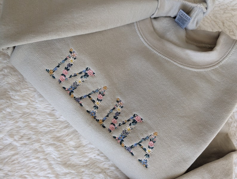 K&ouml;nnte beinhalten: Hellgr&uuml;nes Sweatshirt mit dem gestickten Wort "MAMA" in floralen Buchstaben. Die Buchstaben sind mit rosa Rosen, blauen und gelben Wildblumen und gr&uuml;nen Bl&auml;ttern gef&uuml;llt. Das Sweatshirt ist auf einer wei&szlig;en, strukturierten Oberfl&auml;che gefaltet.