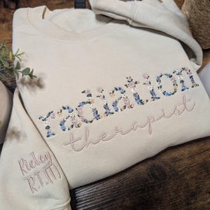 May include: A beige sweatshirt with the words "radiation therapist" embroidered in floral and pink thread. The sleeve has the name "Rieley R.T.(T)" embroidered in pink. A matching folded hoodie is visible in the background.