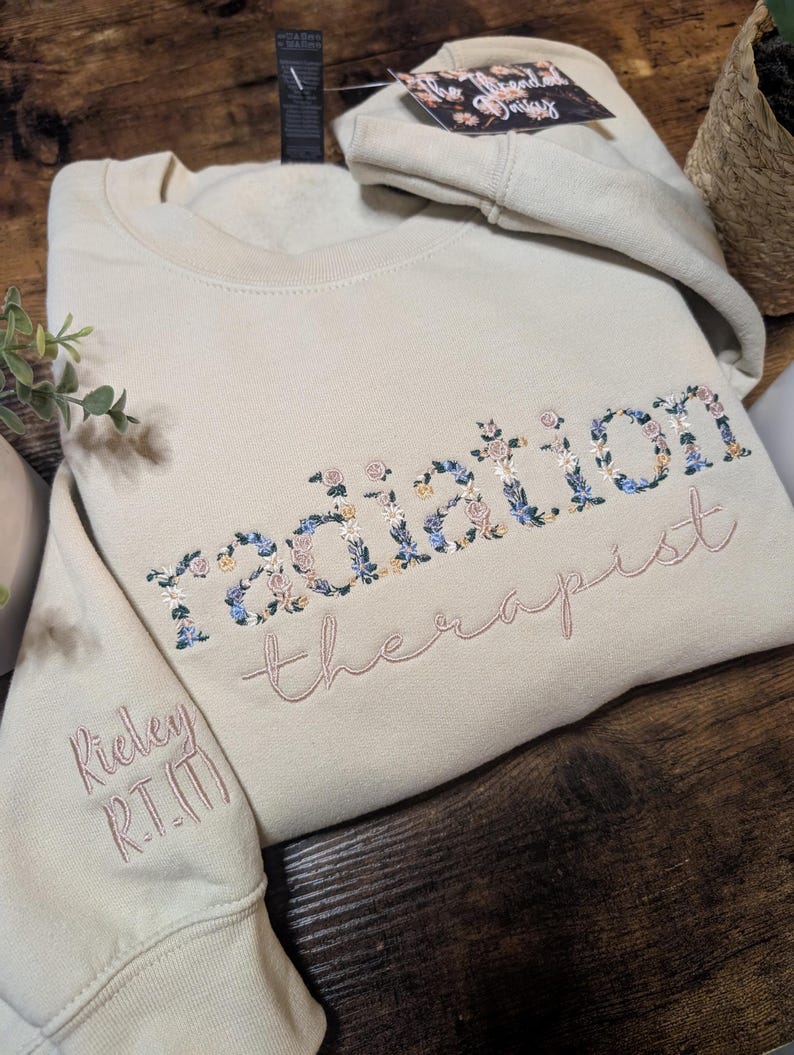 May include: A beige sweatshirt with the words "radiation therapist" embroidered in floral and pink thread. The sleeve has the name "Rieley R.T.(T)" embroidered in pink. A matching folded hoodie is visible in the background.