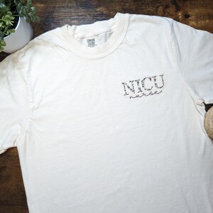 K&ouml;nnte beinhalten: Wei&szlig;es T-Shirt mit einem floralen Design, auf dem vorne "NICU nurse" steht.