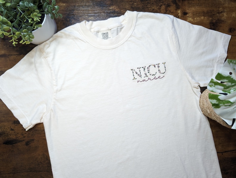 K&ouml;nnte beinhalten: Wei&szlig;es T-Shirt mit einem floralen Design, auf dem vorne "NICU nurse" steht.