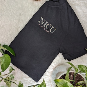 K&ouml;nnte beinhalten: Schwarzes T-Shirt mit den Worten "NICU nurse" in floraler Schrift bestickt.