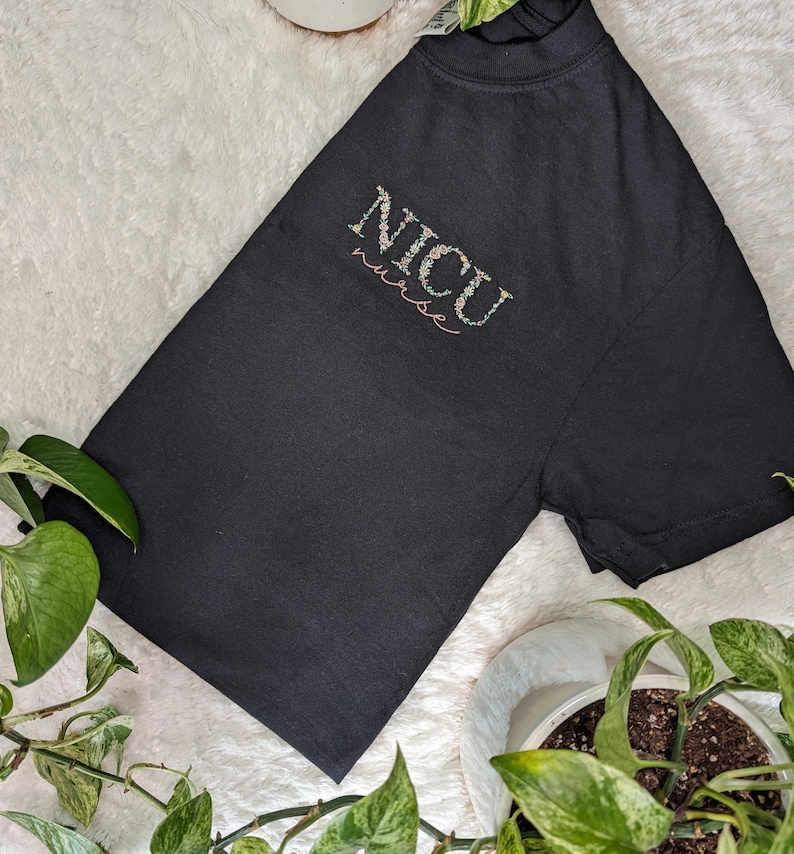 K&ouml;nnte beinhalten: Schwarzes T-Shirt mit den Worten "NICU nurse" in floraler Schrift bestickt.