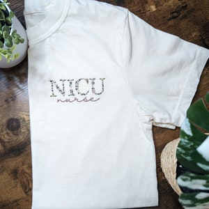 K&ouml;nnte beinhalten: Ein wei&szlig;es T-Shirt mit einem floralen Design, das "NICU nurse" in Rosa buchstabiert.