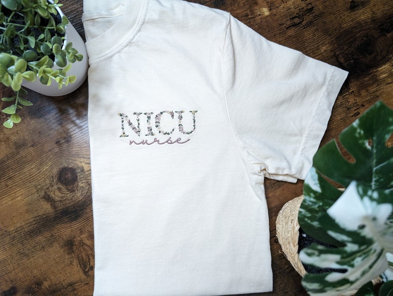 K&ouml;nnte beinhalten: Ein wei&szlig;es T-Shirt mit einem floralen Design, das "NICU nurse" in Rosa buchstabiert.