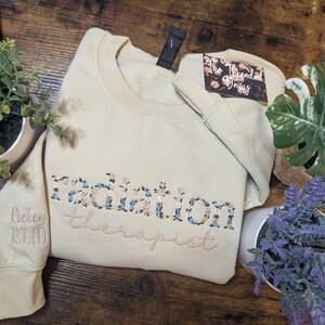 May include: Beige sweatshirt with floral embroidery spelling "radiation therapist." The word "radiation" is filled with blue, pink, and yellow flowers. The sleeve has the name "Ridley R.T.(T)" embroidered in pink.