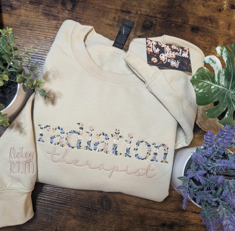 May include: Beige sweatshirt with floral embroidery spelling "radiation therapist." The word "radiation" is filled with blue, pink, and yellow flowers. The sleeve has the name "Ridley R.T.(T)" embroidered in pink.