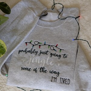 Pu&ograve; includere: Felpa grigio chiaro con la scritta ricamata "probably just going to jingle some of the way I'M TIRED". Il design include una stringa di luci natalizie colorate. Una stringa di luci &egrave; drappeggiata attorno alla felpa.