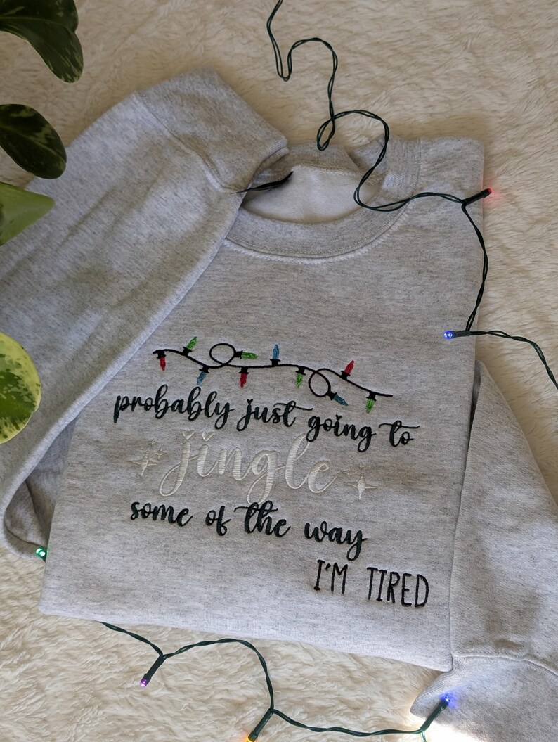 Pu&ograve; includere: Felpa grigio chiaro con la scritta ricamata "probably just going to jingle some of the way I'M TIRED". Il design include una stringa di luci natalizie colorate. Una stringa di luci &egrave; drappeggiata attorno alla felpa.