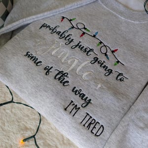 Pu&ograve; includere: Una felpa grigio chiaro con testo ricamato e una fila di luci natalizie. Il testo recita "probably just going to Jingle some of the way I'M TIRED" in filo nero e bianco. Le luci natalizie sono rosse, verdi e blu.