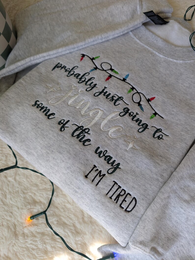 Pu&ograve; includere: Una felpa grigio chiaro con testo ricamato e una fila di luci natalizie. Il testo recita "probably just going to Jingle some of the way I'M TIRED" in filo nero e bianco. Le luci natalizie sono rosse, verdi e blu.