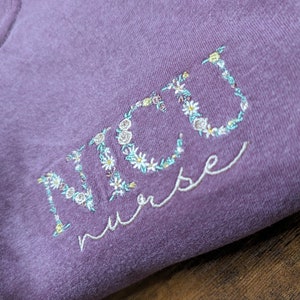 K&ouml;nnte beinhalten: Lilafarbenes T-Shirt mit dem Wort "NICU nurse" in wei&szlig; gestickt mit floralen Akzenten.
