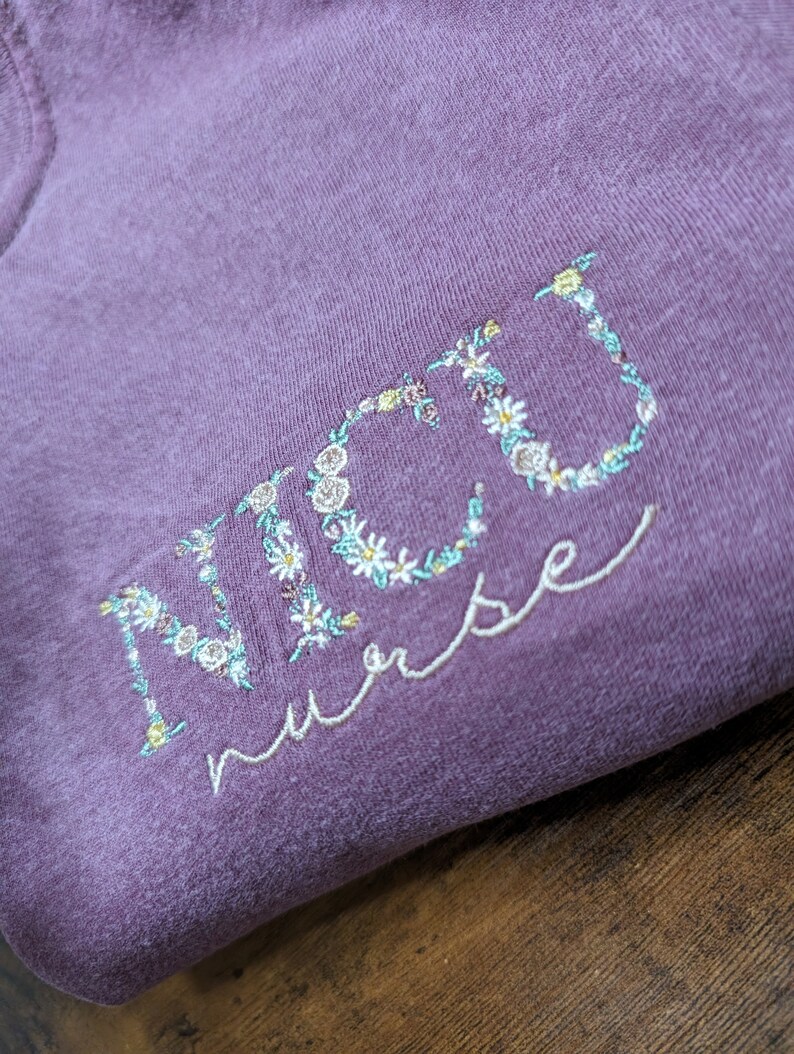 K&ouml;nnte beinhalten: Lilafarbenes T-Shirt mit dem Wort "NICU nurse" in wei&szlig; gestickt mit floralen Akzenten.