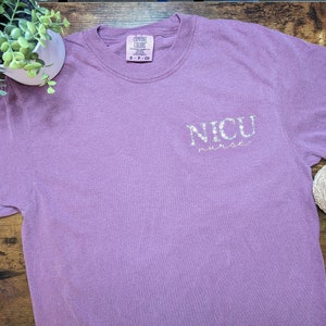 K&ouml;nnte beinhalten: Ein lila T-Shirt mit dem Schriftzug "NICU nurse" in wei&szlig;en Glitzerbuchstaben.