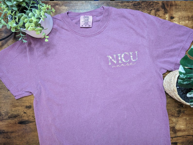 K&ouml;nnte beinhalten: Ein lila T-Shirt mit dem Schriftzug "NICU nurse" in wei&szlig;en Glitzerbuchstaben.